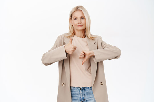 Image of mature blond woman, stylish senior lady shows thumbs up and thumbs down, like or dislike, standing over white background