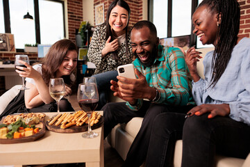 Multi ethnic bunch young adults guffawing at african american man smart phone during friends house party, drinking wine, smiling, laughing, happily enjoying mates gathering.