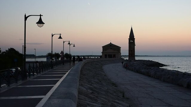 People stroll at dawn on the seafront of Caorle, in the background the sanctuary "Madonna dell'Angelo" built on a promontory surrounded by rocks.