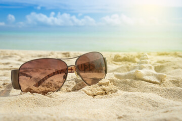 Sunglasses on the background of a blurred sea and sky. Sunglasses and a seashell on a sunny beach. The concept of vacation, relaxation and sunbathing.