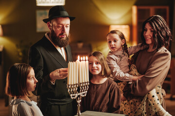 Portrait of happy jewish family lighting menorah candle during Hanukkah celebration in cozy home - obrazy, fototapety, plakaty
