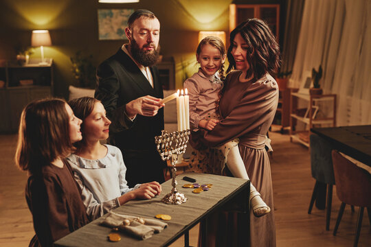 Portrait of modern jewish family lighting silver menorah candle during Hanukkah celebration in cozy home setting