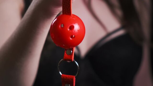 Young girl in black lingerie close-up holds a red bdsm gag on a black background. BDSM is an outfit for adult sex games.
