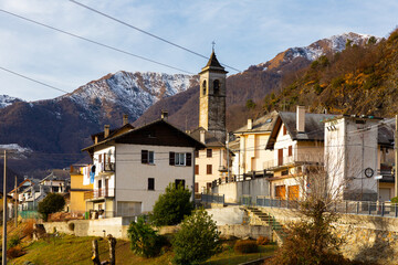 Fototapeta na wymiar View of residential houses and bell tower of parish church of small Alpine township of Finero located in Cannobina valley on background of snowy mountain ridge on winter day, Piedmont, Italy..