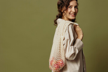 Smiling curly cute female in linen casual shirt hold string bag with fruit behind her back looks at...