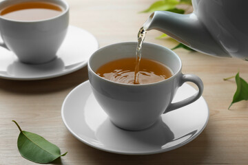 Pouring green tea into white cup with saucer and leaves on wooden table, closeup