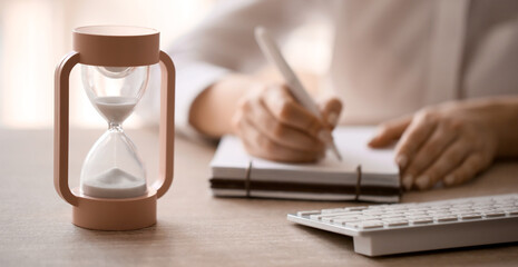 Hourglass on table and woman writing something in notebook. Deadline concept