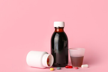 Bottle of cough syrup, pills and cup on pink background