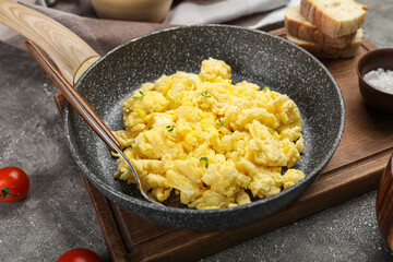 Wooden board with frying pan of tasty scrambled eggs on grunge background, closeup