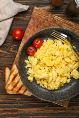 Frying pan with tasty scrambled eggs, tomatoes and toasts on wooden table, closeup