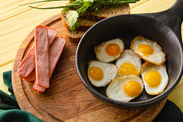 Board with frying pan of tasty fried quail eggs and sausages on color wooden background, closeup