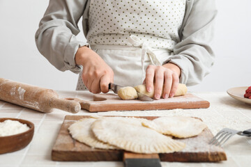 Woman cutting dough for chebureks at table near light wall, closeup