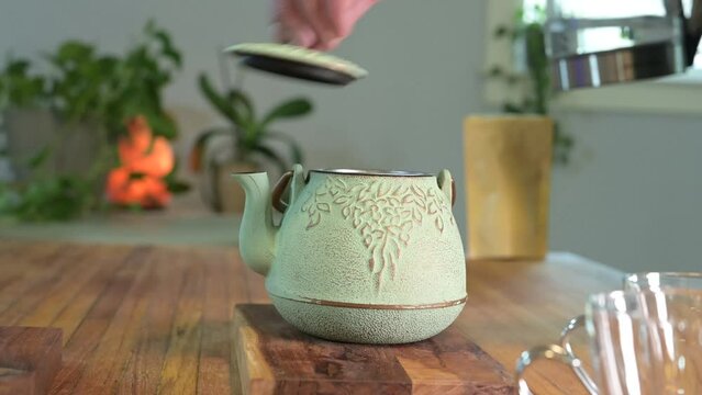 Making herbal tea in a green teapot on a wood table in a light and airy room with green plants in the background and a glass teacup