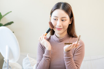 Happy routine beauty concept, pretty asian young woman, girl make up face by applying powder foundation by brush around face, looking at the mirror at home. Female look with natural fashion style.
