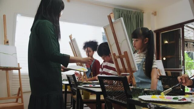 A group of multiracial kids learning with a female Asian teaches acrylic color picture painting on canvas in art classroom, creatively learning with skill at the elementary school studio education.