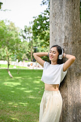 Calm Asian female listening to music through her headphones, leaning on the tree