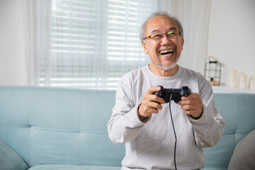 Asian senior old man enjoying holding joystick playing video game at home in living room, mature man hands using game controller, Funny on retirement elderly smile sitting on sofa life gaming © sorapop