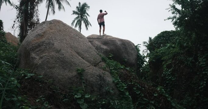 View from the back of young athletic man tourist in pink shirt and casual shorts running up on a rock, contemplates an exotic rainforest nature. He raises his arms up and breathes fresh mountain air