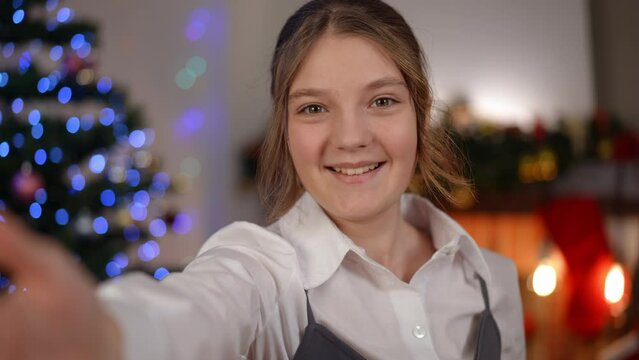 Selfie video portrait of charming teenage girl looking at camera standing at background of Christmas lights at home. Happy positive relaxed Caucasian teenager filming blog posing indoors on holiday
