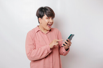 Fototapeta premium Surprised short-haired Asian woman wearing a pink shirt pointing at her smartphone, isolated by a white background