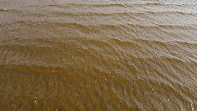 A still shot from a drone down into the clear sea water with small ripples. A dark orange sandy bottom is visible through the layer of water. Slowness and regularity of time in nature
