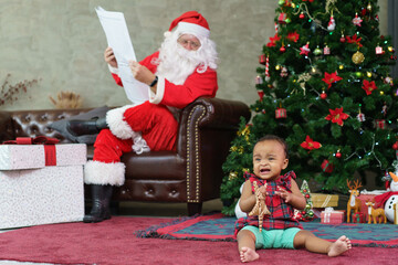 Fototapeta na wymiar Senior man in Santa Claus costume reading newspaper on sofa watching African American baby girl playing and crying on the floor. Christmas holiday season