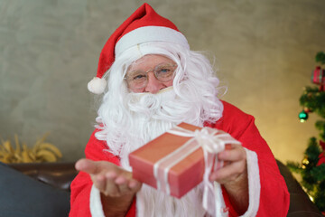 Senior man wearing eyeglasses in Santa Claus costume open hand giving present box to camera. Christmas holiday