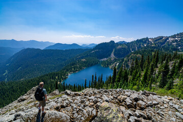 Fototapeta na wymiar Adventurous athletic man standing on a mountain top overlooking an alpine lake and a rugged mountain range on a beautiful sunny day in the Pacific Northwest.