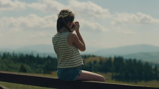 Young photographer shooting landscape on beautifull green hillway using digital camera, sitting on wooden fence, taking pictures of nature. Girl filling the portfolio with amazing photos.