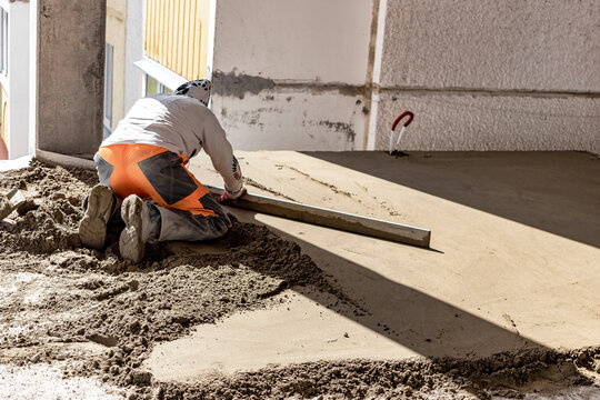 Close up of man builder placing screed rail on the floor covered with sand-cement mix at construction site. Male worker leveling surface with straight edge while screeding floor. Blurred background.