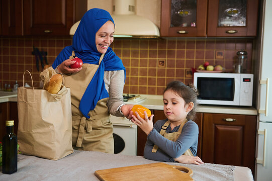 Middle-Eastern Muslim Cute Woman In Hijab, Puts A Bag Of Groceries On The Table. Little Girl Helps Her Loving Mom Unpacking Shopping Bags With Healthy Food In The Kitchen. Eco-friendly Paper Packaging