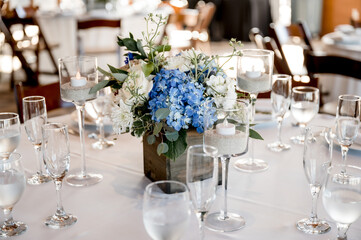 Table with blue hydrangeas and white roses wedding floral centerpiece on white table cloth surrounded by place settings and glassware