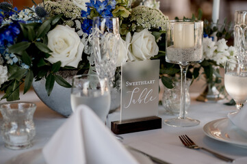 Sweetheart sign on table with blue and white wedding floral centerpiece on white table cloth surrounded by place settings and glassware. blue hydrangeas white roses horizontal no people