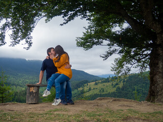 Young man and woman admiring breathtaking view while sitting on bench in the mountains. Lovely couple in casual clothes sit under large tree with forest background.
