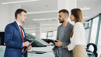 Interested young couple man and woman are talking to manager in car dealership while he is telling them about luxury auto model. Beautiful people, transportation and business concept.