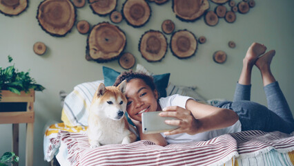 Cheerful girl is taking selfie with cute pet posing with beautiful dog lying on bed having fun and laughing. Modern apartment with lovely design and furniture is visible. - 539372148