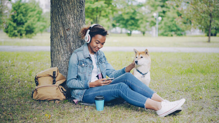 Modern African American girl is listening to music with wireless headphones and using smartphone relaxing in city park with pet dog, stroking and caressing the animal.