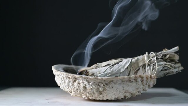 Burning sage in a shell on a wood table with black background showing sage smoke