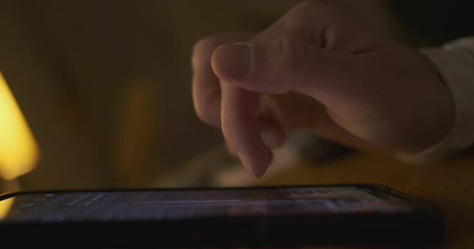Man starts flipping through a feed of pictures on a smartphone in a dark room, doomscrolling. The phone is on the table, a close-up of the index finger.
