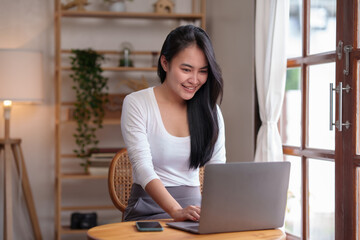 Bright, cute and happy Asian female student study online in the living room using laptop computer in a relaxed manner. Young Asian woman using laptop at home.