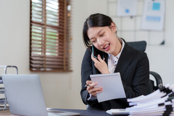 Happy Asian businesswoman smiling and talking on the phone with a client in the office with a bunch of papers on the desk.
