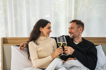 European couple smiling looking at each other in winter clothing holding glasses of champagne sitting on bed under blanket.