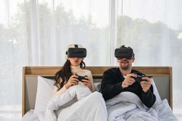 Caucasian couple wearing a VR headsets goggle device and holding game controller sitting on their bed playing game together.