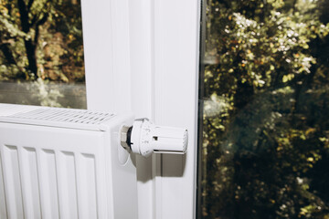 White metal battery at home near a panoramic window with a thermostat close-up. Apartment heating