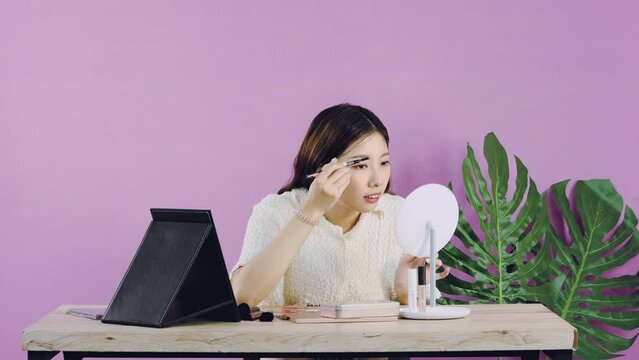 Young asian woman in white shirt long hair sitting and drawing eyebrow and make up while recording her vlog using lablet camera on pink color background wall. Video 4K.
