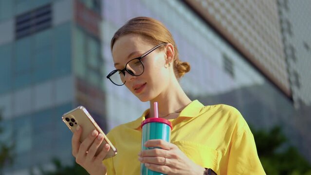 Businesswoman Using Phone Outdoors - orbit shot Close-up. One Attractive Business Woman in Glasses Holding Tumbler and Checking Emails on Mobile Phone Outside Office Building
