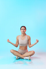 Fototapeta na wymiar Young woman practicing her meditation, sitting in a yoga pose on a mat, teal studio background
