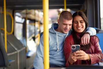 Couple sitting on a bus seat, looking at a phone, hugging and having good times browsing the...