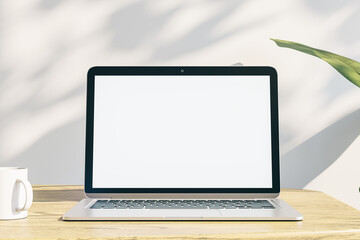  designer office desktop with empty white laptop display with mock up place in frame, concrete wall background with sunlight and shadows, blurry decorative plant and coffee cup. 3D Rendering.