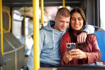 Couple sitting on a bus seat, looking at a phone, hugging and having good times browsing the internet while commuting - 539405574